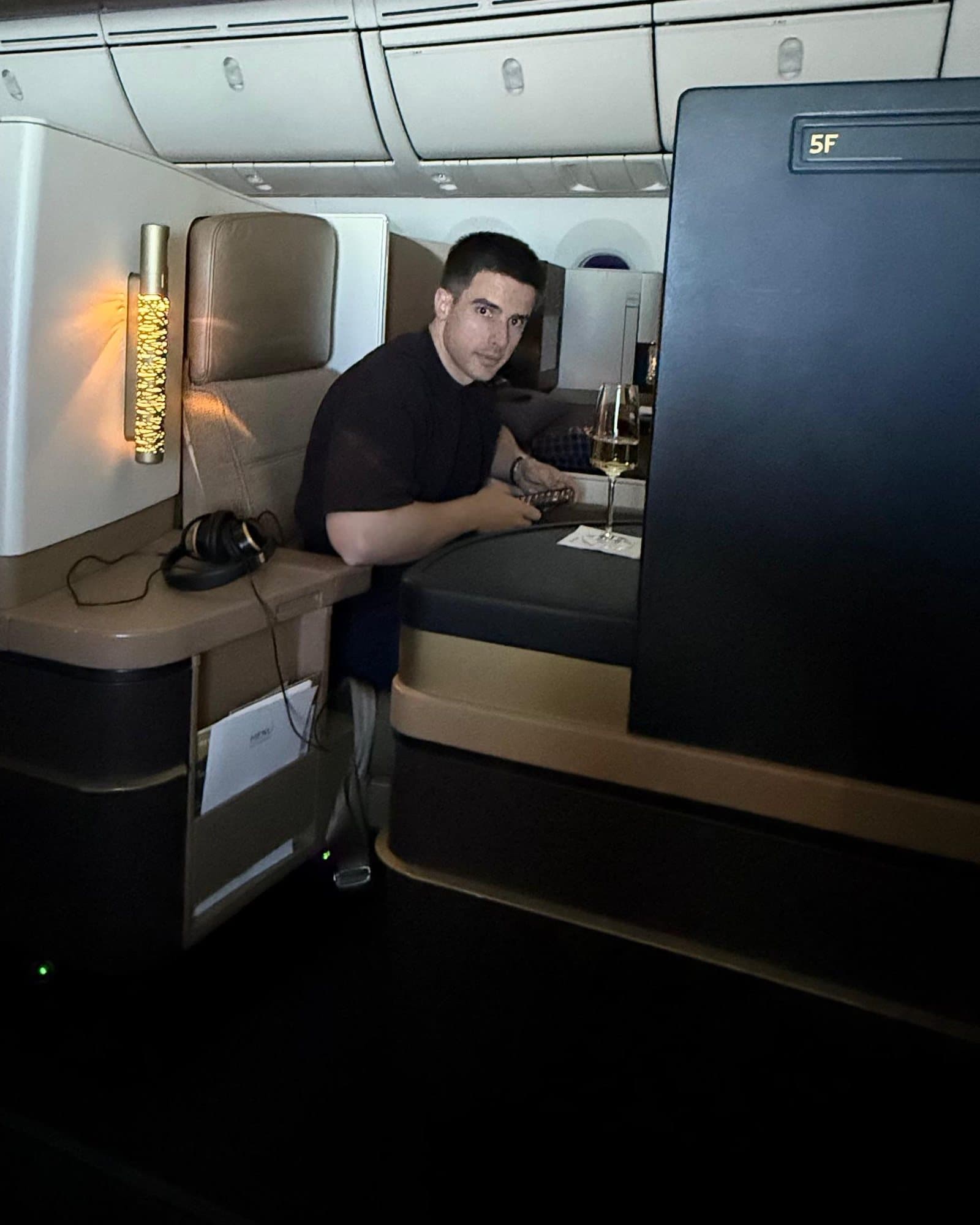 Ramón in seat 5F of a plane, looking up from a phone, a glass of white wine on the side table.